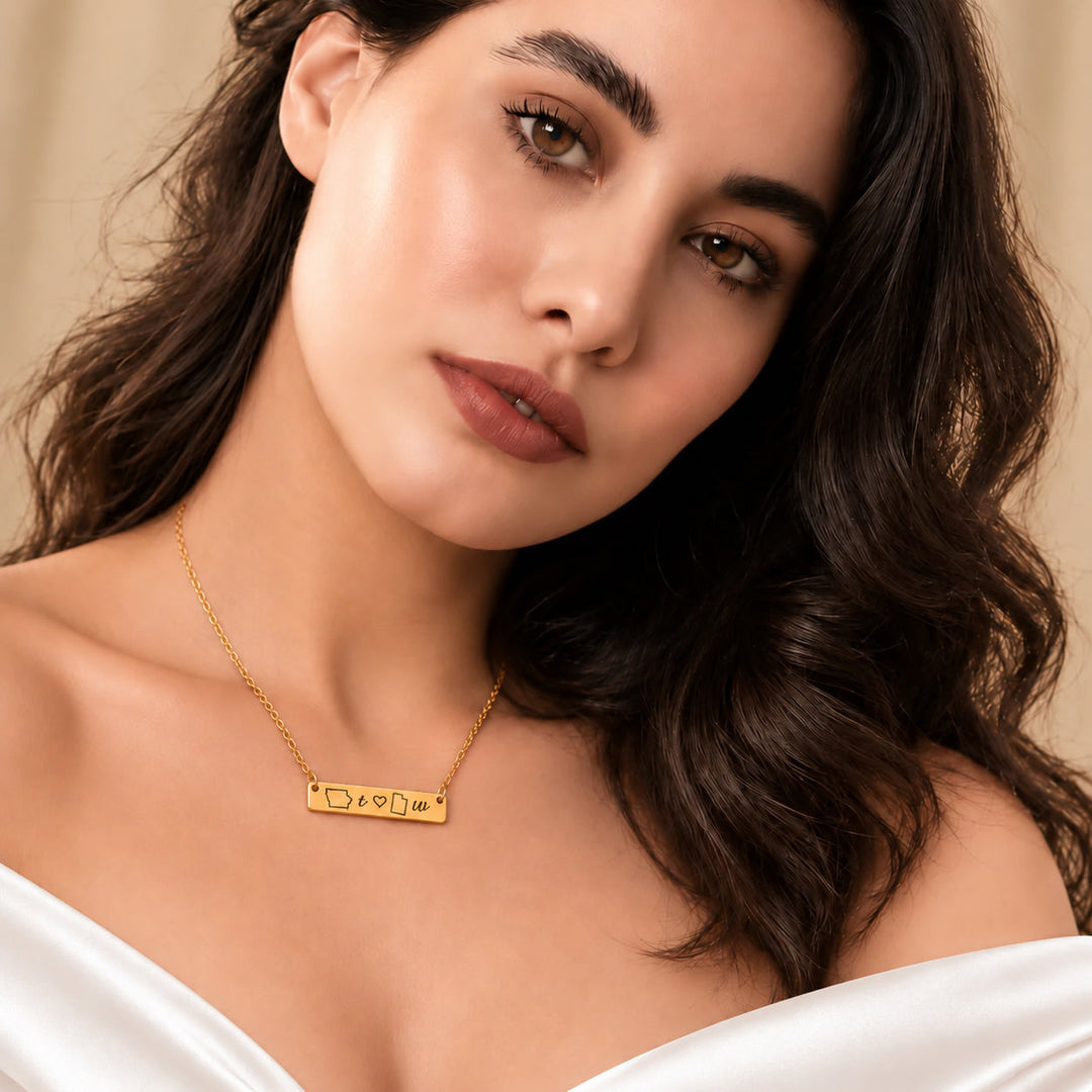 Woman in a white off-shoulder top wearing a gold bar necklace engraved with two state outlines, two lowercase initials and a small heart