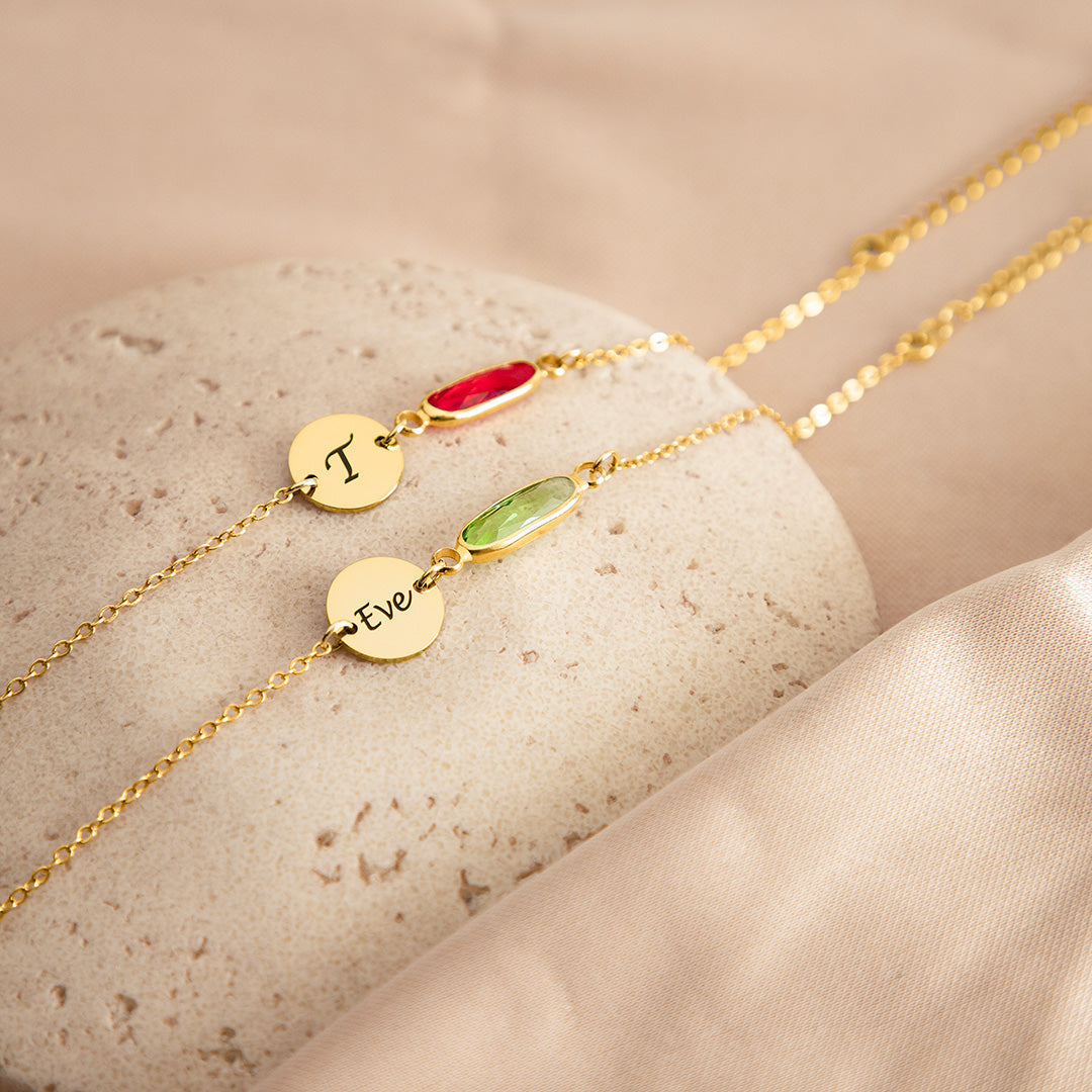 Two VIVO Minimalist Birthstone Initial Bracelets in gold vermeil displayed on a stone surface showing initial T with red stone and Eve with green stone