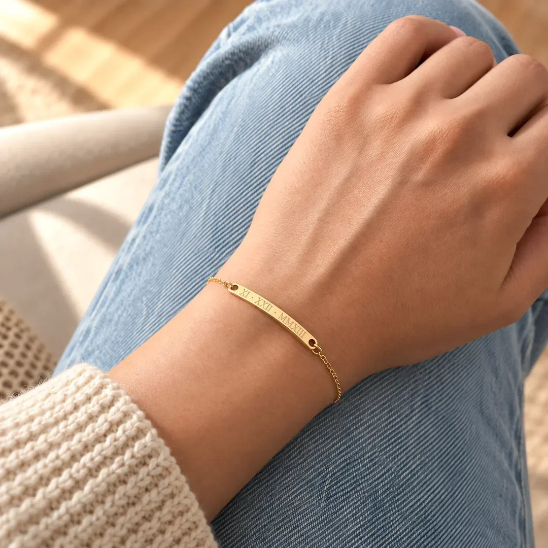 Gold vermeil name bar bracelet with a Roman numeral date, worn on the wrist with denim and a knit sleeve