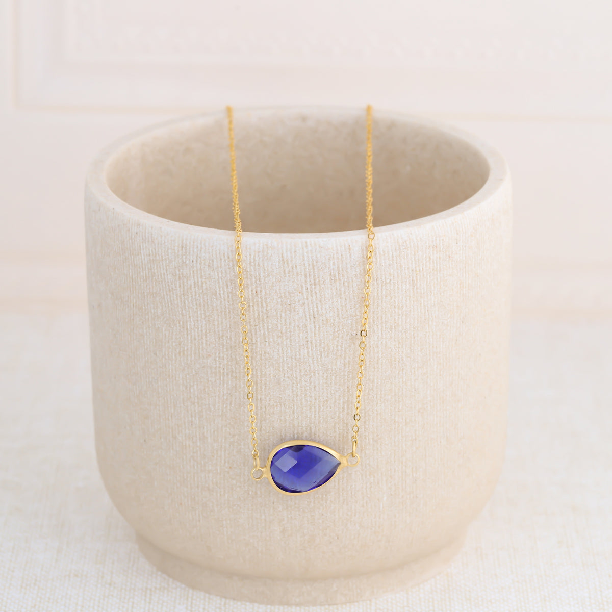 Gold-tone necklace with a dark blue faceted teardrop pendant displayed on a beige ceramic cup