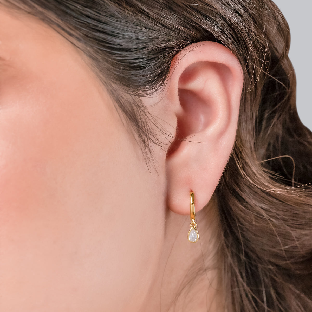 Gold-tone dangle huggie hoop earring with a clear teardrop stone worn on the ear against a neutral background