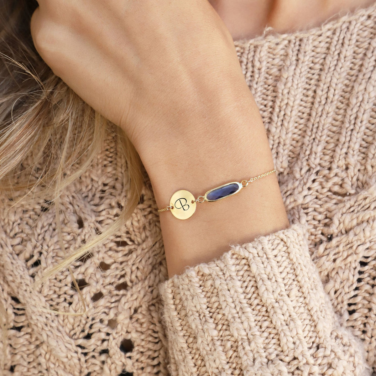 Gold-tone bracelet with engraved B initial disc and purple elongated stone worn on a wrist against a beige knit sweater