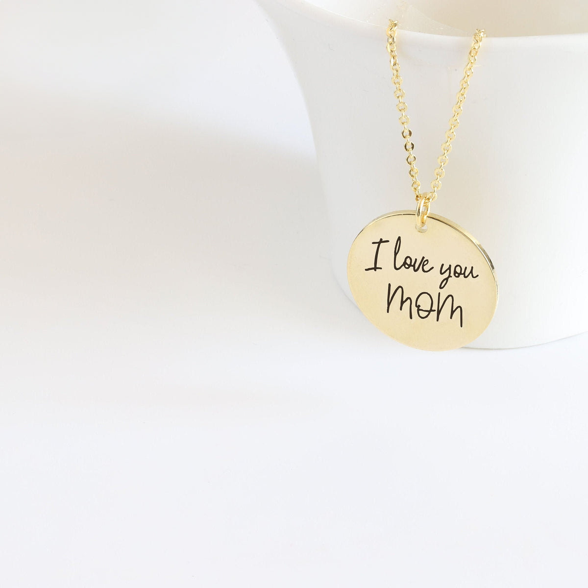 Gold-tone round pendant necklace engraved I love you Mom in handwritten script displayed against a white curved stand