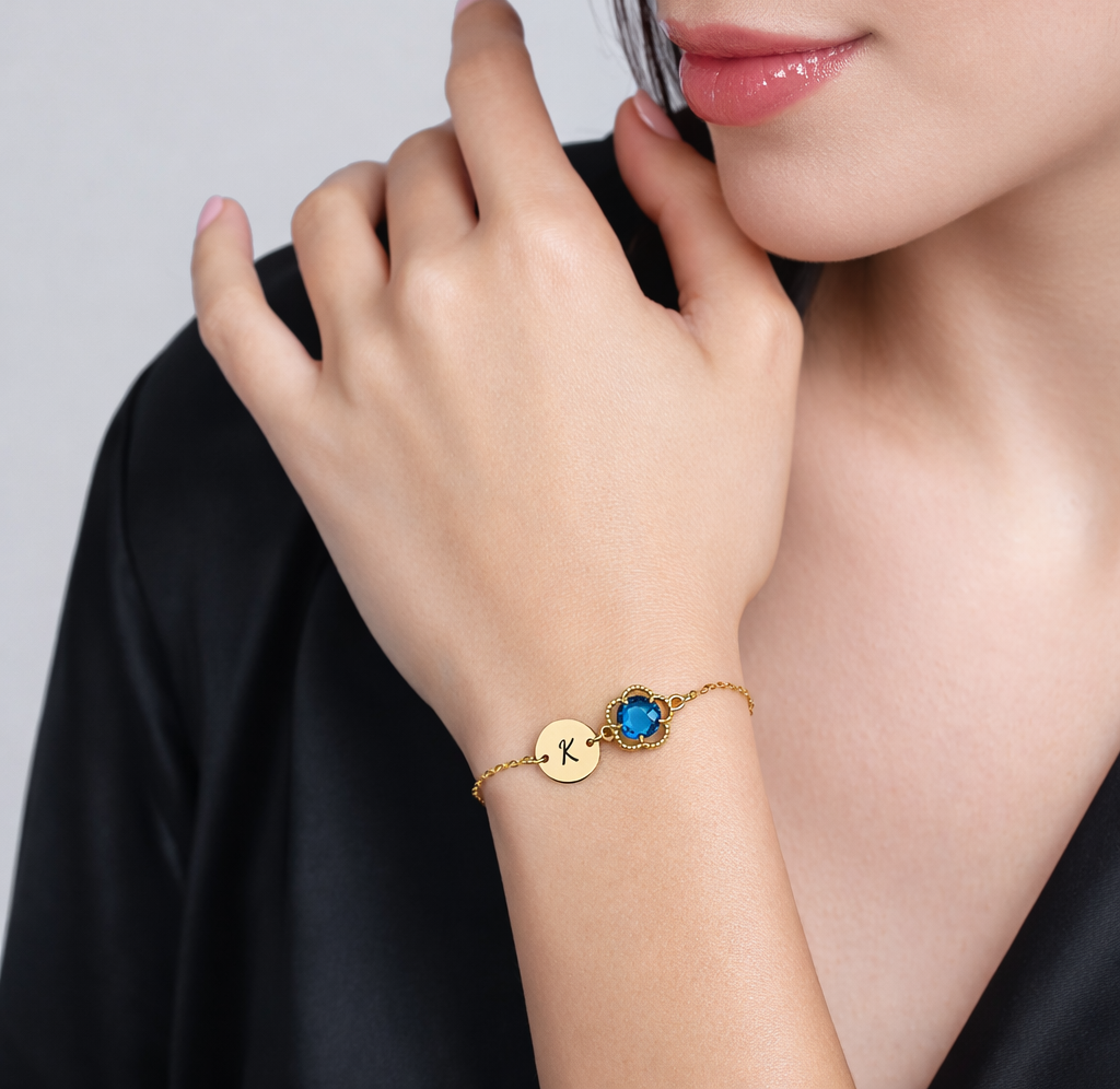 Gold-tone bracelet with engraved K initial disc and blue round stone in a beaded frame worn on a wrist