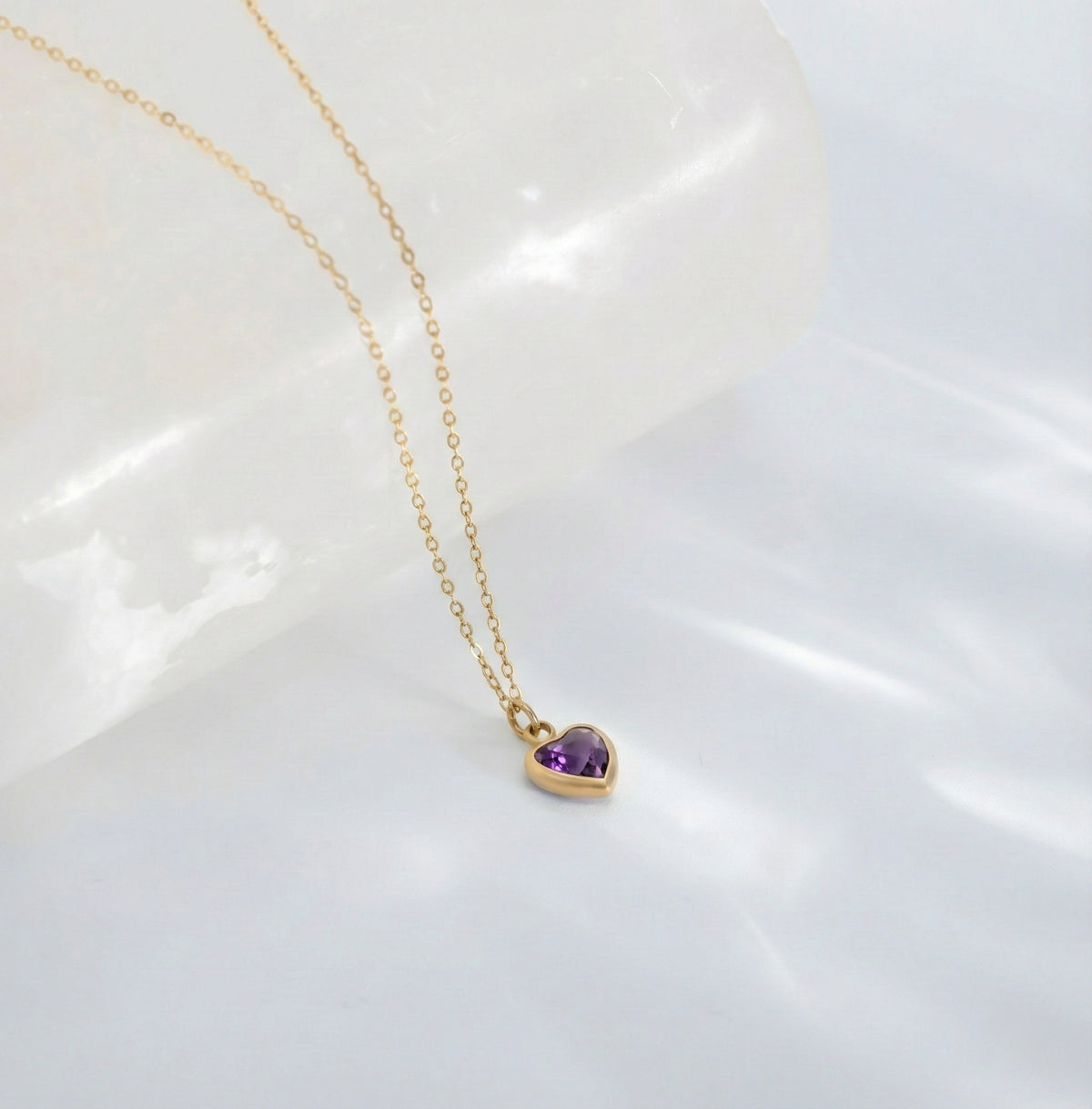 Gold-tone necklace with a purple heart-shaped stone pendant displayed on a white sculptural surface