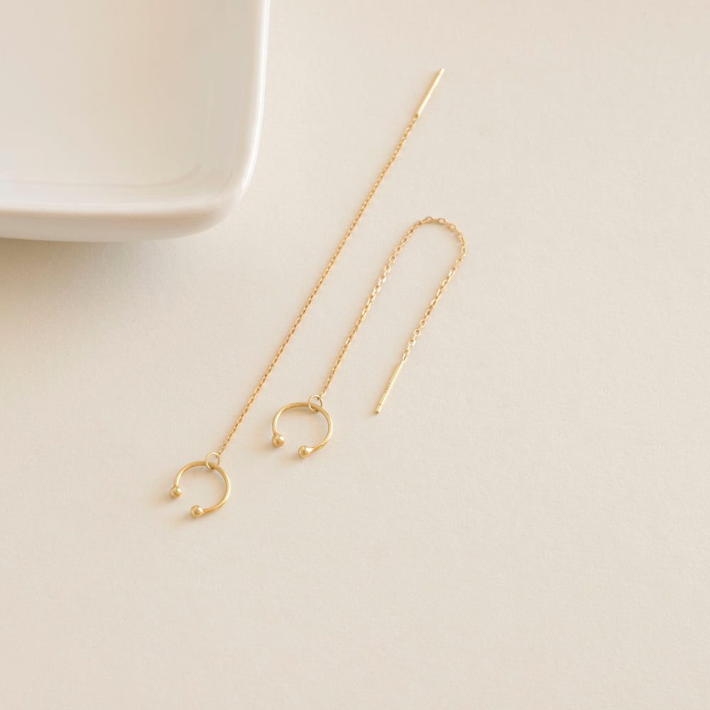Gold chain earrings on a light beige surface with a white dish in the background
