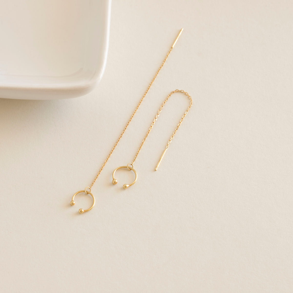 Gold chain earrings on a light beige surface with a white dish in the background