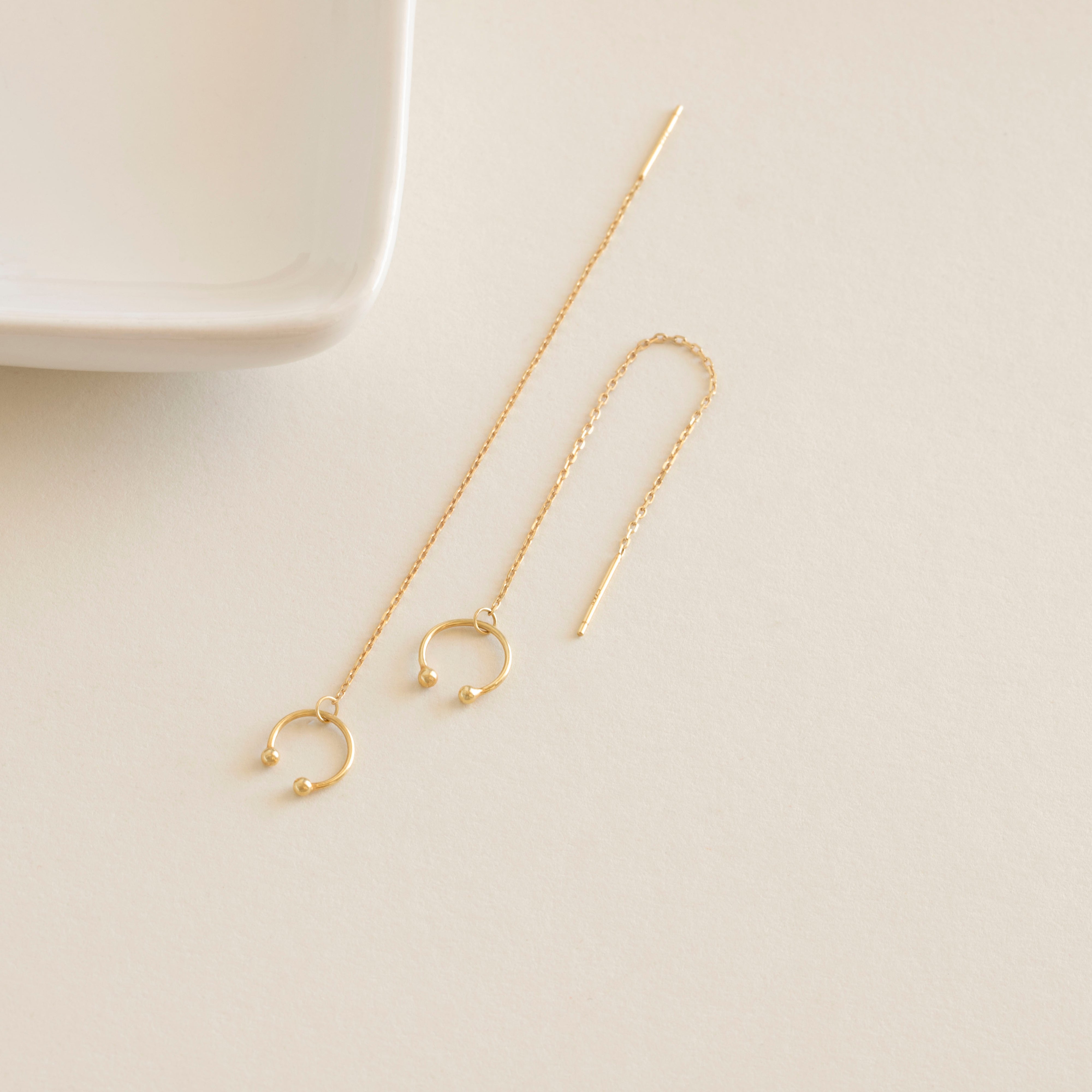 Gold chain earrings on a light beige surface with a white dish in the background
