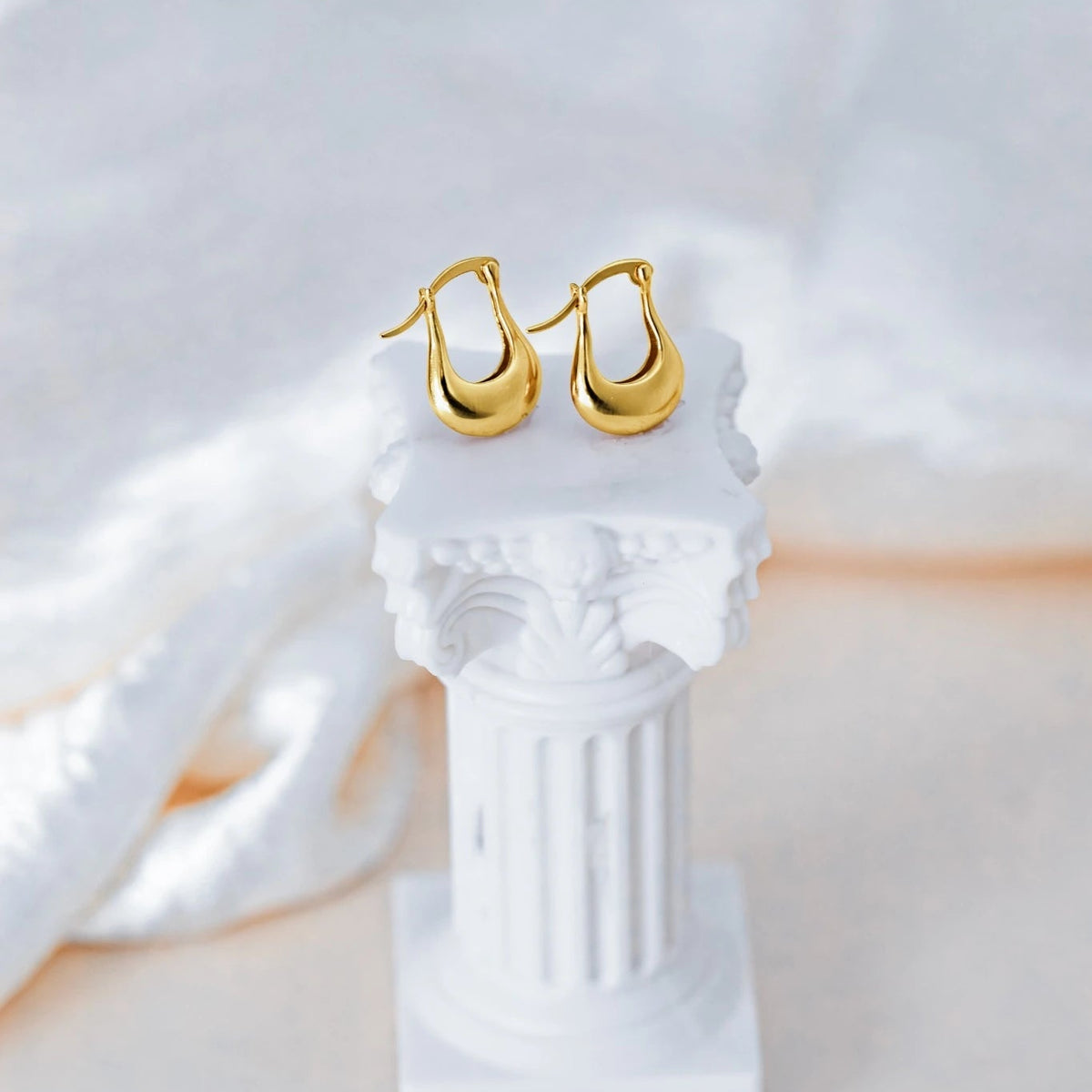 Gold hoop earrings on a white pedestal with a white background