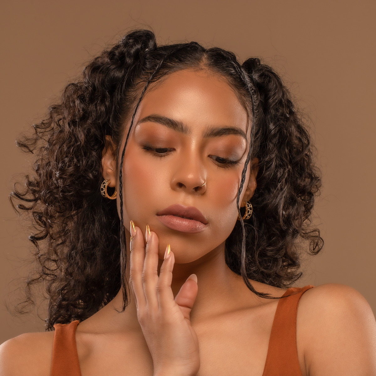 Woman with styled hair and a neutral expression wearing gold checkered hoops against a brown background