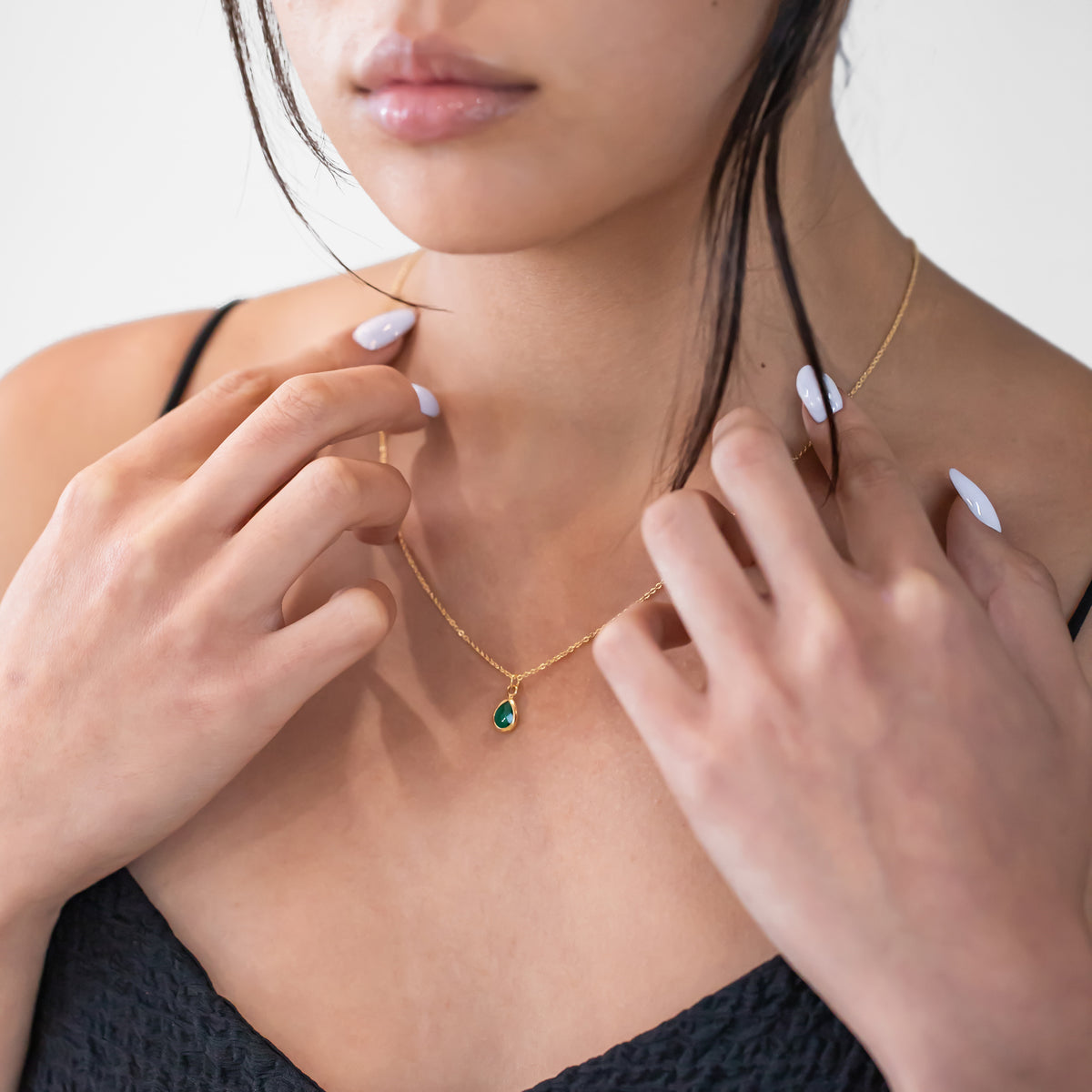 Gold-tone necklace with a green teardrop pendant worn at the neckline with hands near the chain