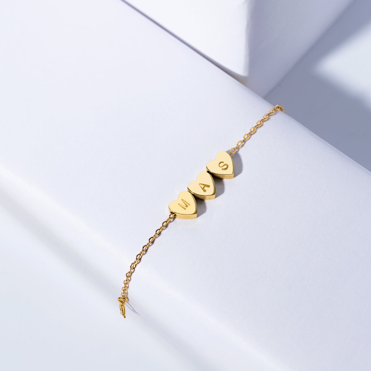 Gold-tone bracelet with three engraved heart charms marked M, A, and S on a white surface