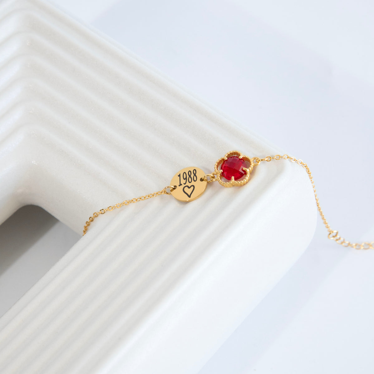 Gold-tone bracelet with round disc engraved 1988 and heart beside a red round stone in a beaded frame on white surface