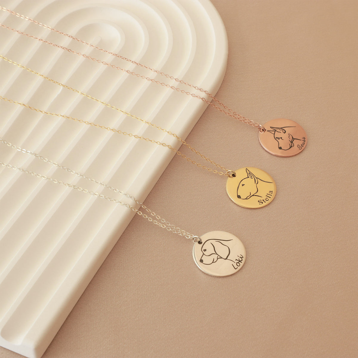 Three round pet silhouette necklaces in rose-gold, gold-tone, and silver-tone finishes on a beige surface