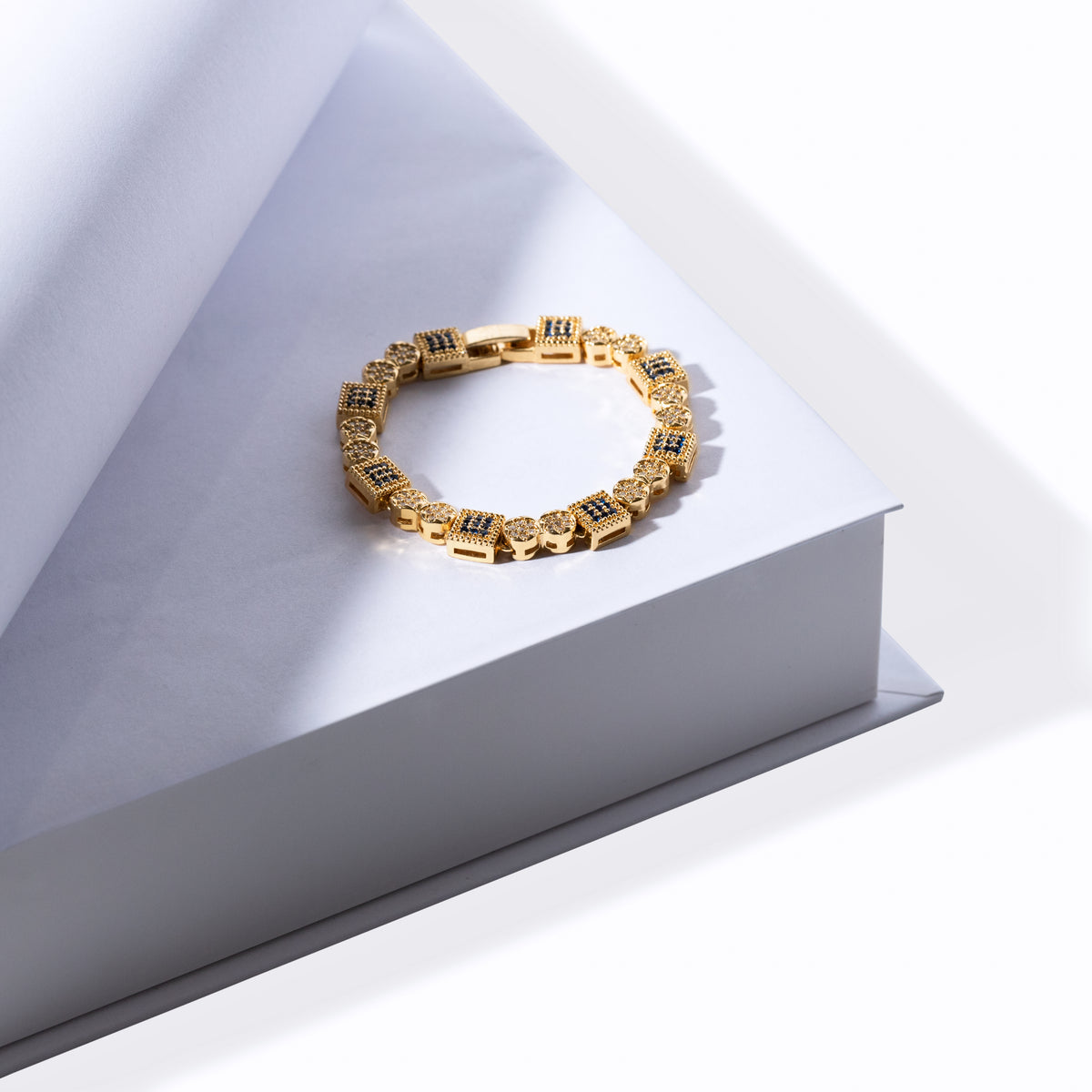 Gold-tone geometric bracelet with square blue accents and round clear accents displayed on a white pedestal
