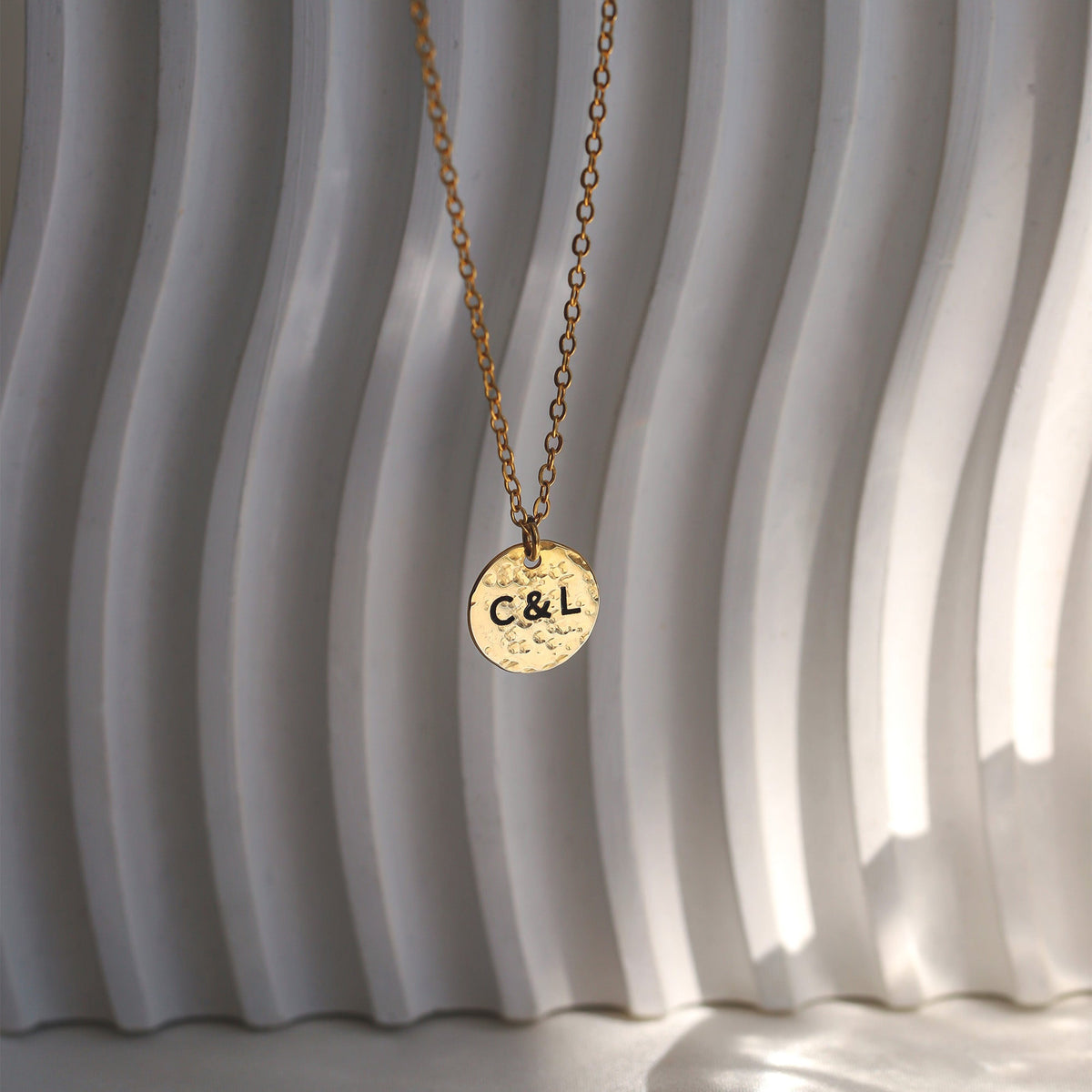Gold-tone hammered disc necklace engraved with C&L against a sculpted white background