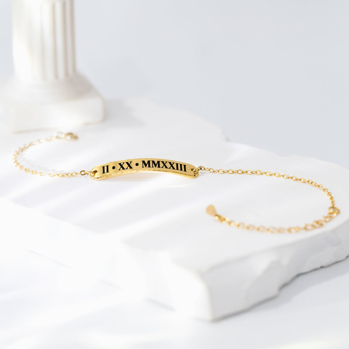 Gold-tone bar bracelet engraved with Roman numerals II XX MMXXIII on a white sculptural surface