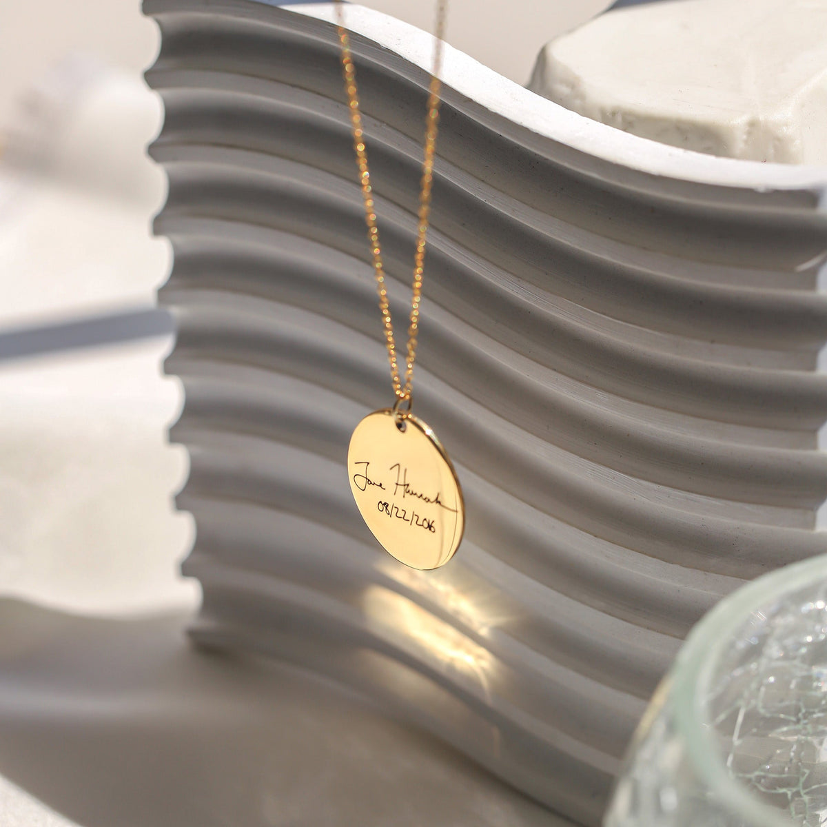 Gold-tone oval necklace with handwritten engraving and a date, displayed against a sculpted gray background