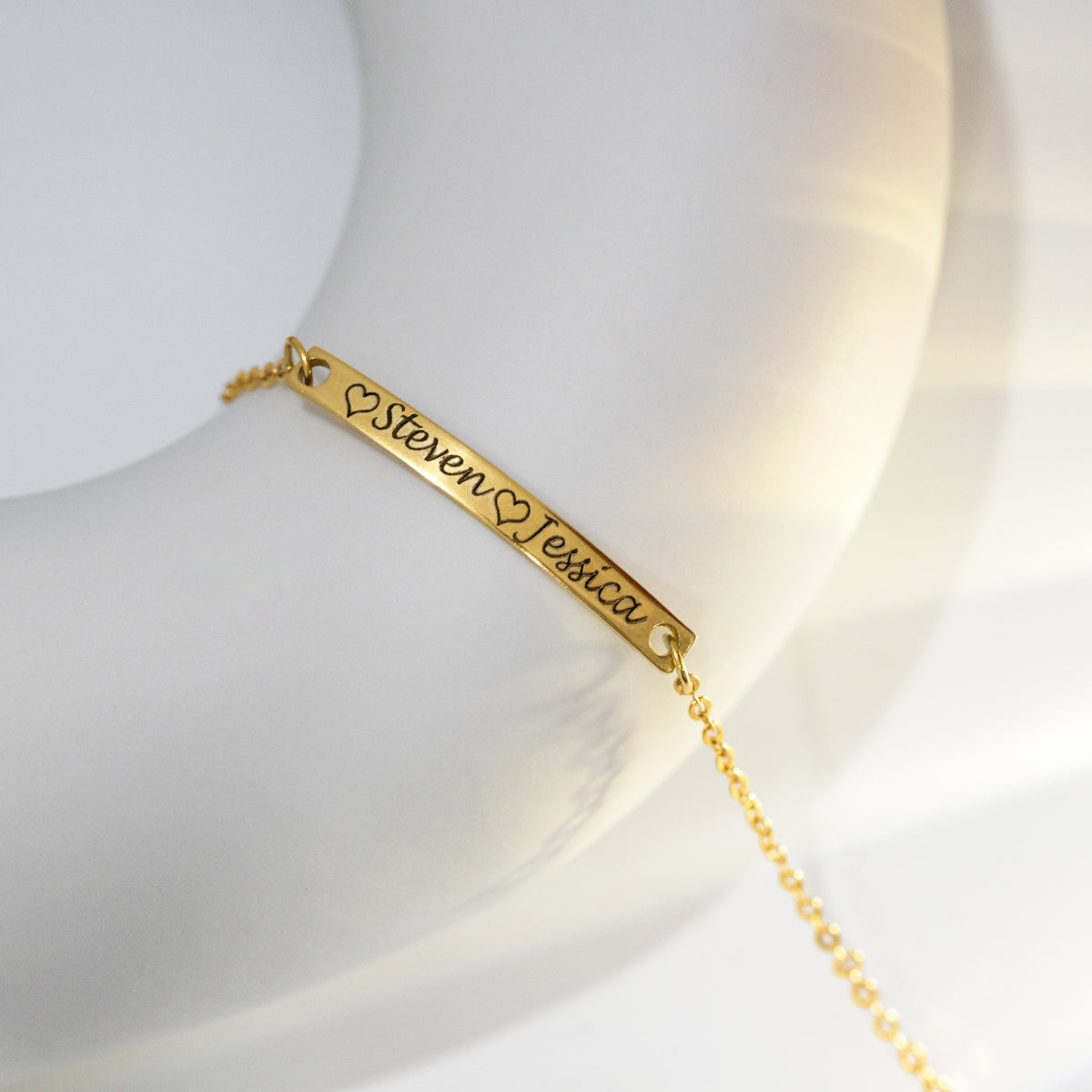 Gold-tone bar bracelet engraved with hearts and the names Steven and Jessica on a white sculptural surface