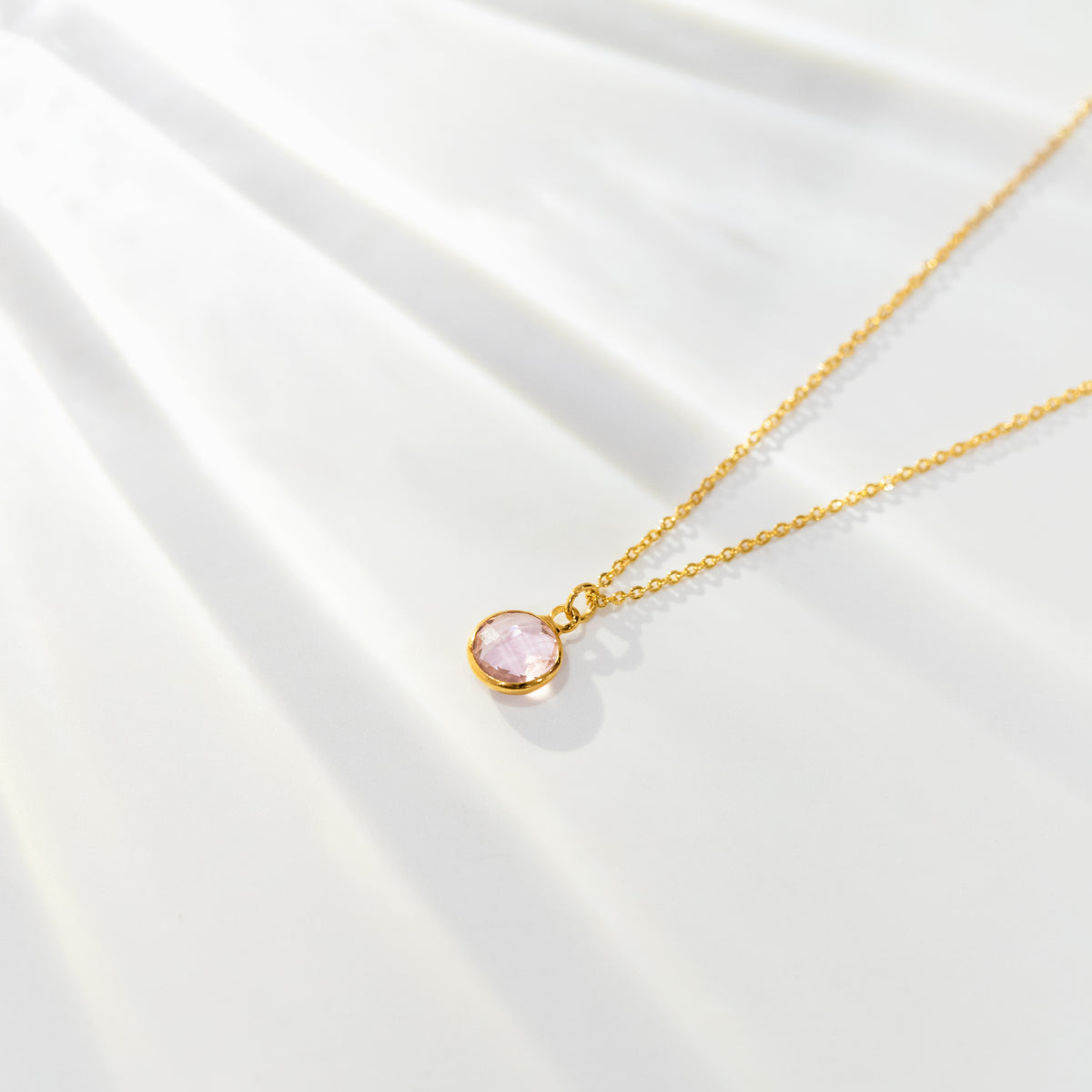 Gold-tone necklace with a round pink stone pendant displayed on softly folded white fabric