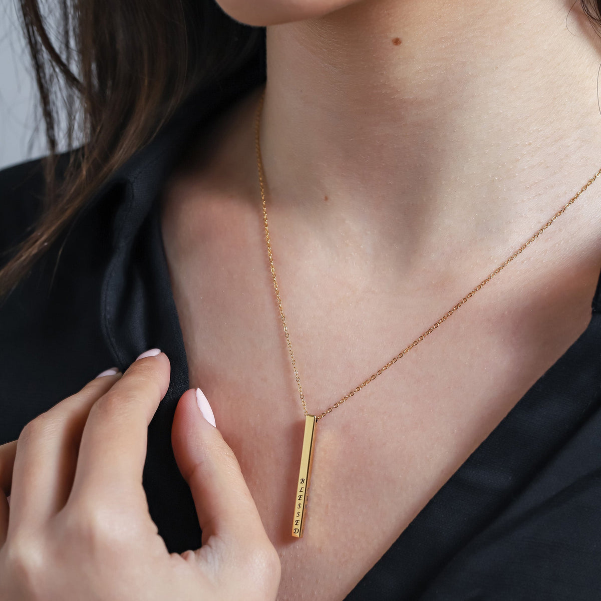 Gold-tone vertical bar necklace engraved BLESSED, worn on the neck against a black top