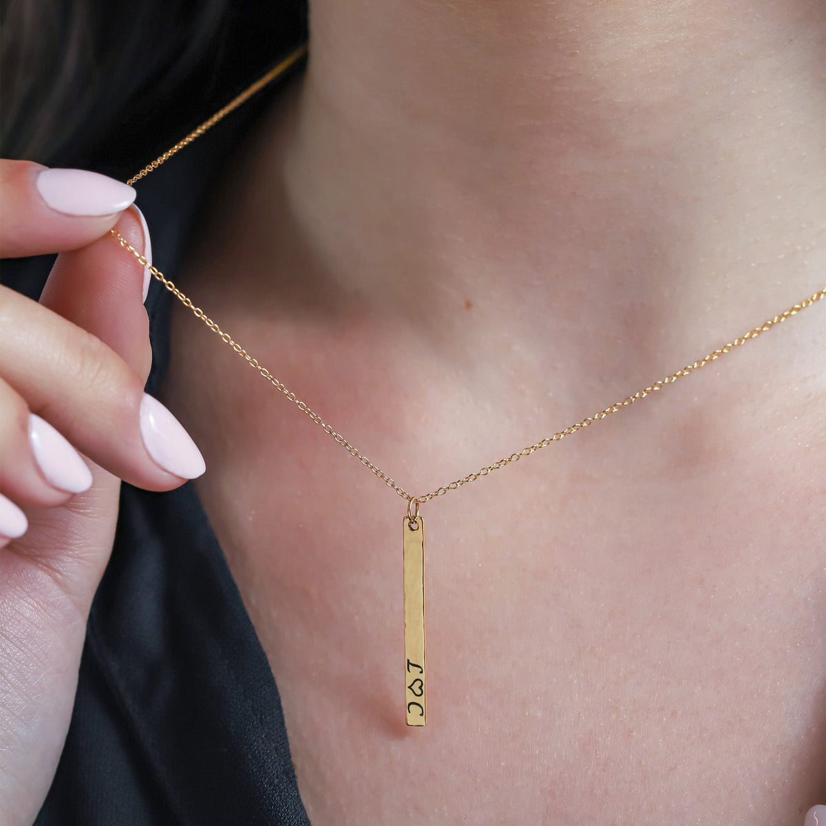Gold-tone vertical bar necklace with small engraved initials and heart detail, worn on the neck