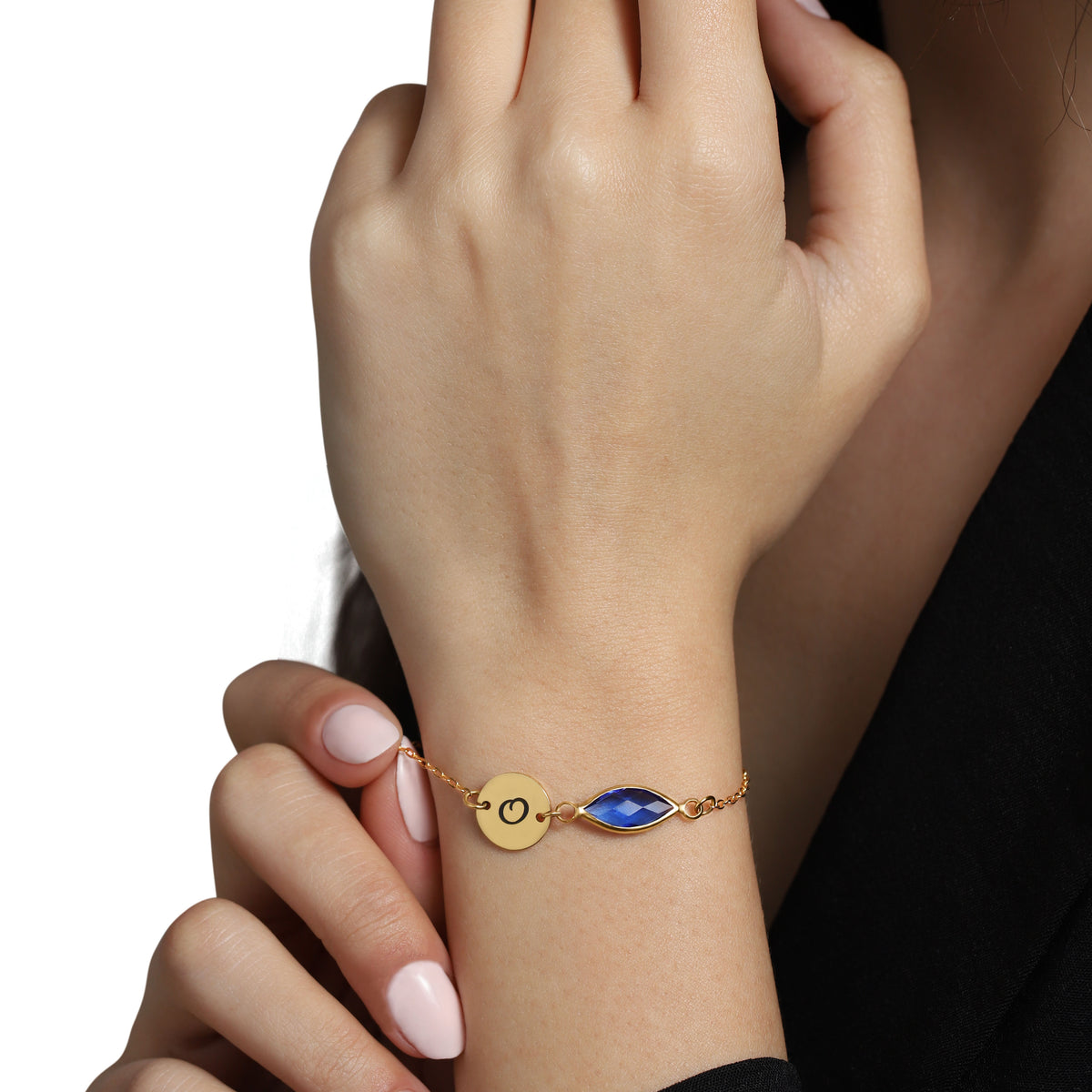 Gold-tone bracelet with engraved O initial disc and blue faceted marquise-shaped stone worn on a wrist
