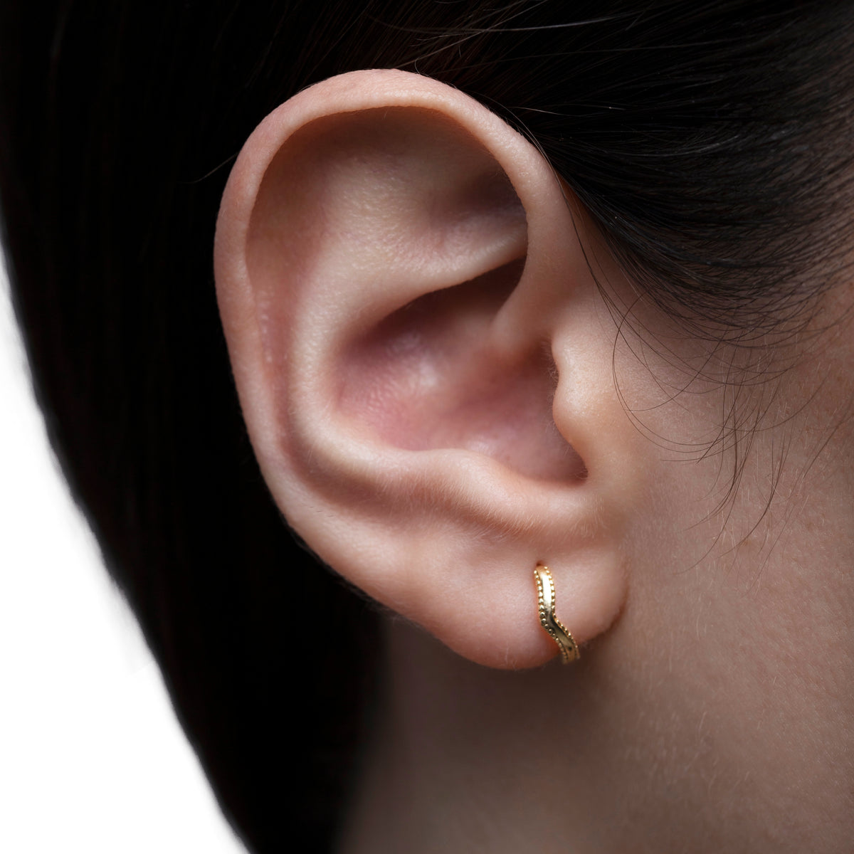 Gold-tone huggie hoop earring with angled textured front worn on the ear in a close-up view