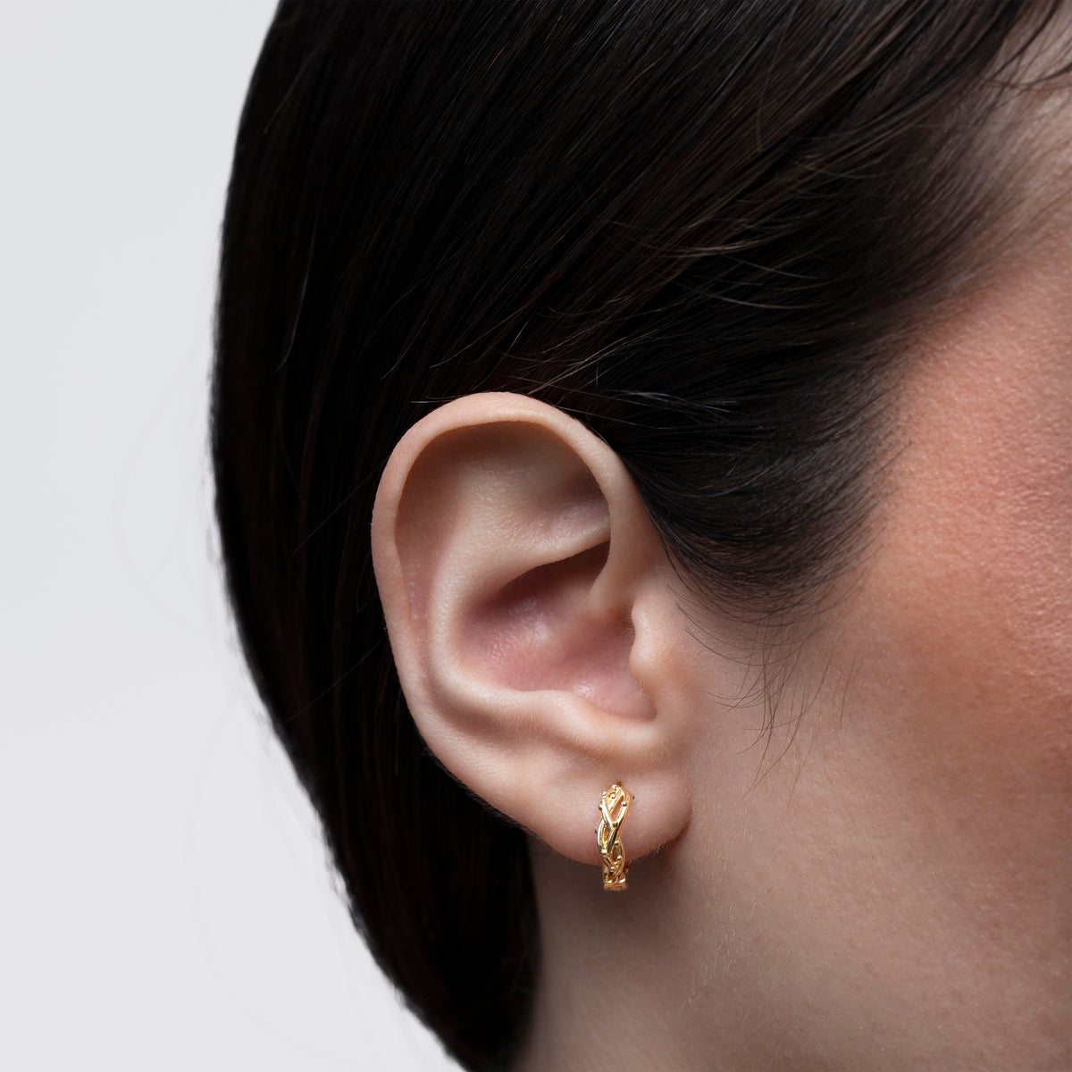 Gold-tone woven huggie hoop earring worn on the ear against a light gray background