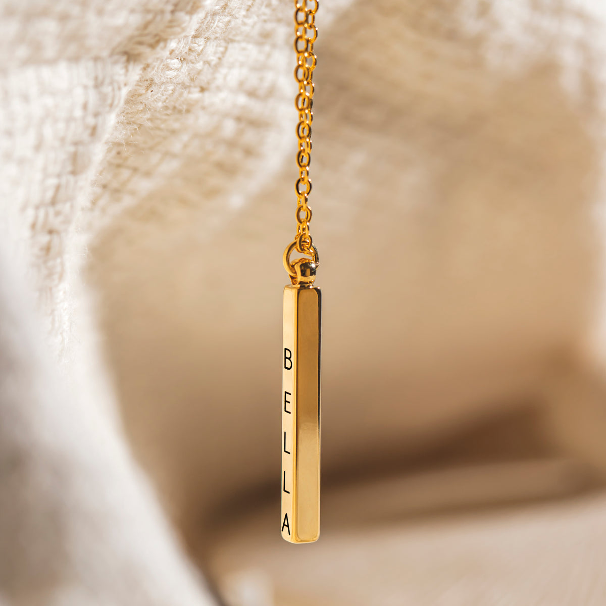 Gold-tone vertical bar pendant necklace engraved BELLA displayed against cream fabric