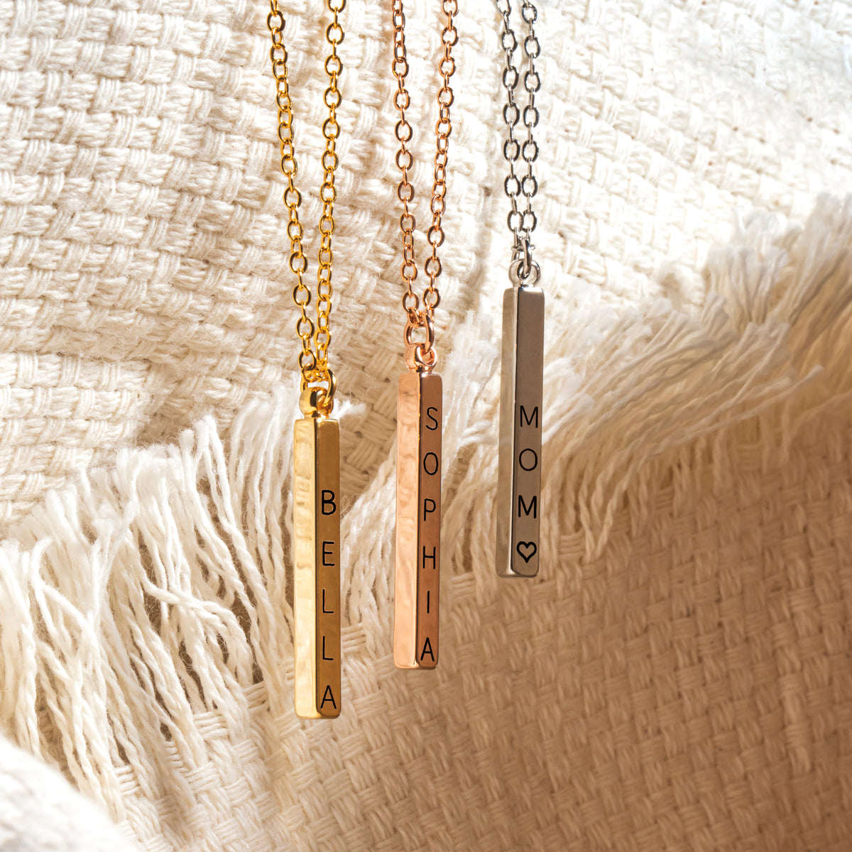 Gold-, rose-gold-, and silver-tone vertical bar necklaces engraved BELLA, SOPHIA, and MOM with a heart on cream fabric