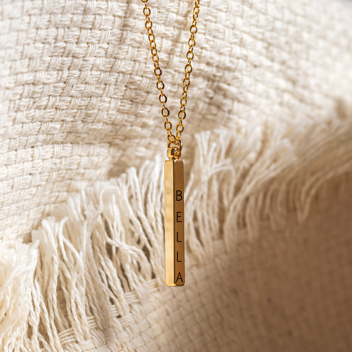 Gold-tone vertical bar pendant necklace engraved BELLA displayed on textured cream woven fabric
