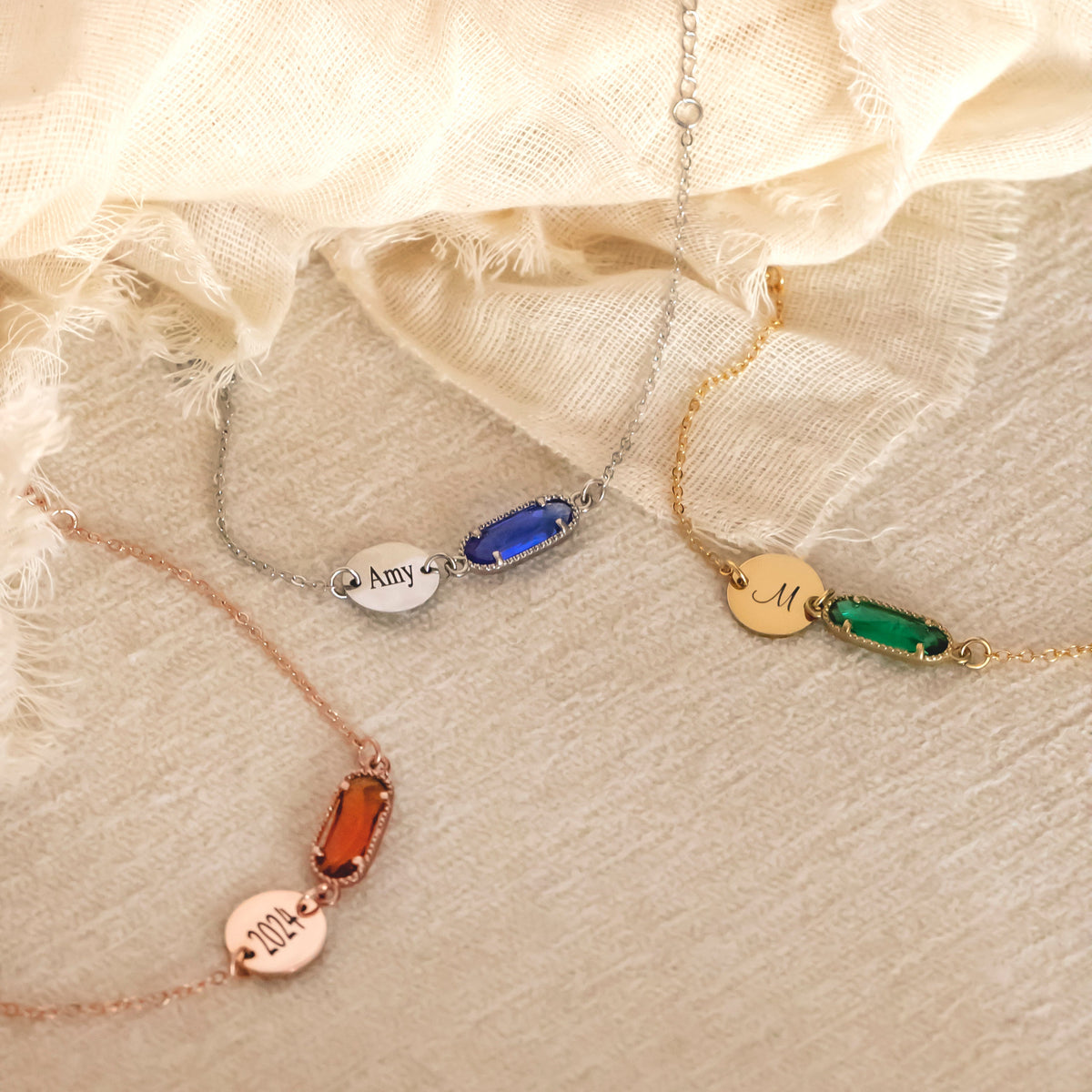 Three bracelets with engraved 2024, Amy, and M discs and orange, blue, and green rectangular stones on beige fabric