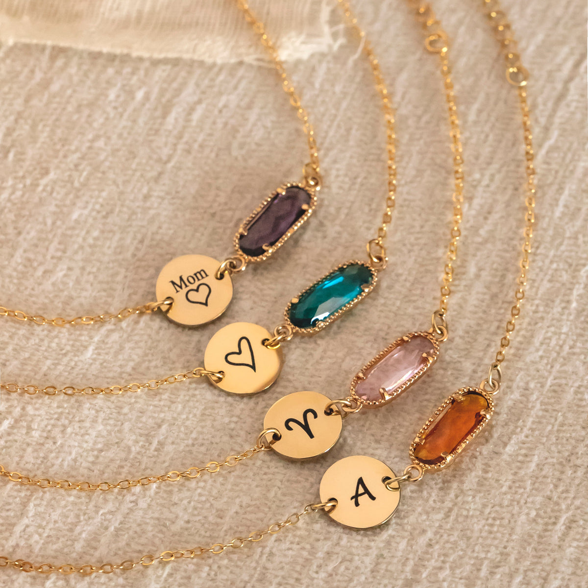 Four bracelets with engraved round discs and purple, green, pink, and amber rectangular stones on beige fabric