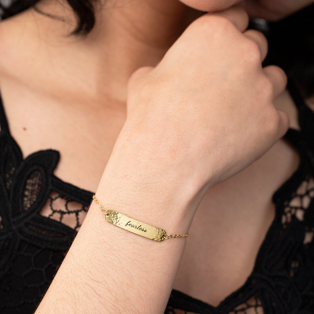 Gold bracelet with 'fearless' engraving on a wrist, worn by a person in a black lace garment.