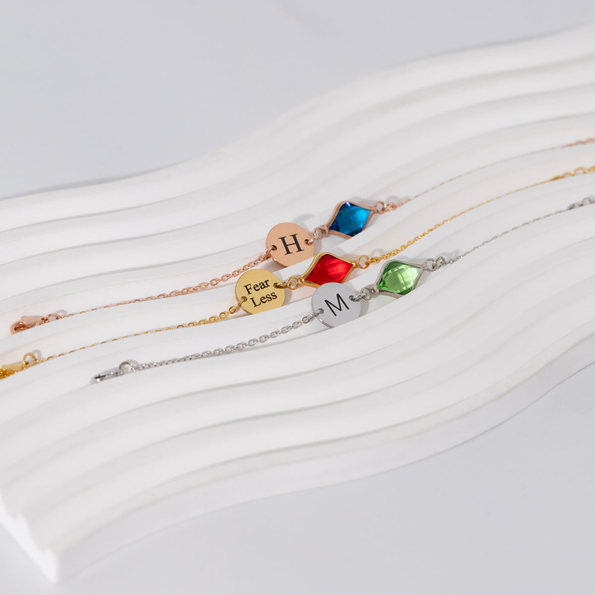 Three bracelets with engraved H, Fear Less, and M discs and blue, red, and green faceted stones on white ribbed surface