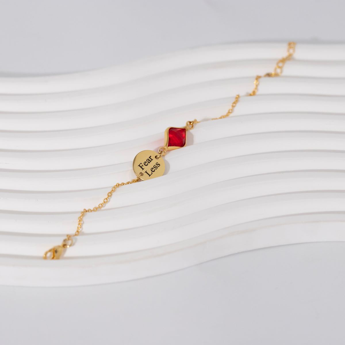 Gold-tone bracelet with round disc engraved Fear Less and red faceted rectangular stone on a white ribbed surface