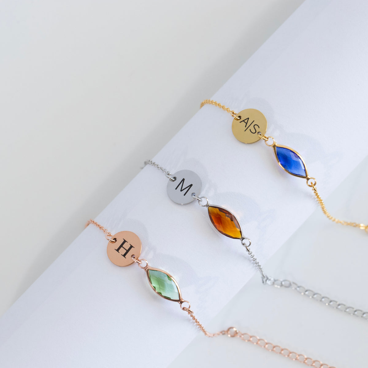 Three bracelets with engraved round discs and blue, amber, and light green marquise-shaped stones on white surface