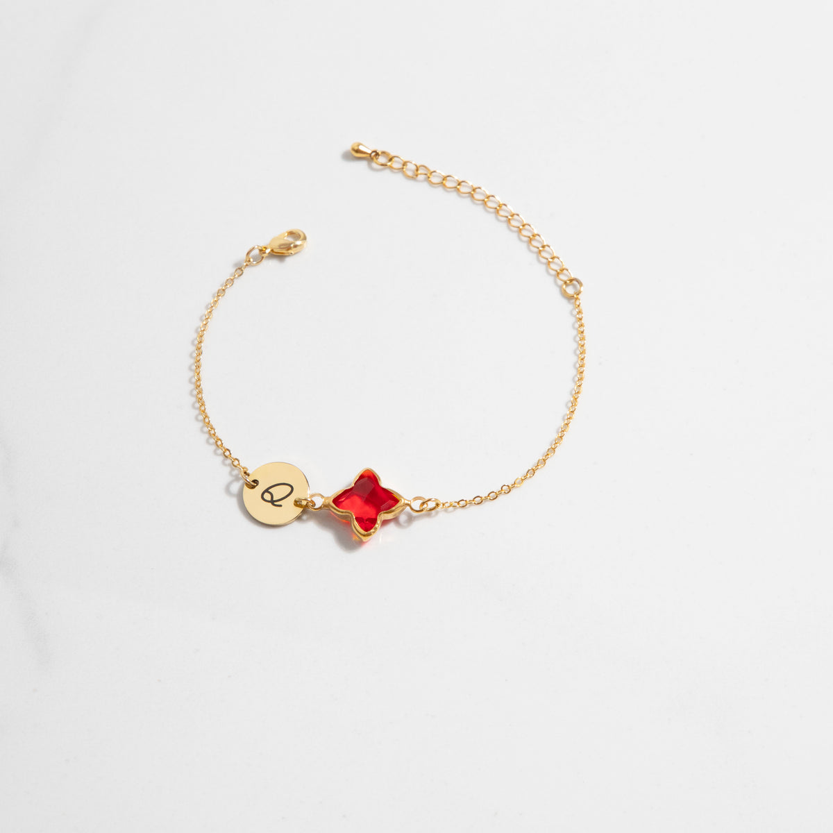 Gold-tone bracelet with engraved Q initial disc and red four-lobed stone on a light background