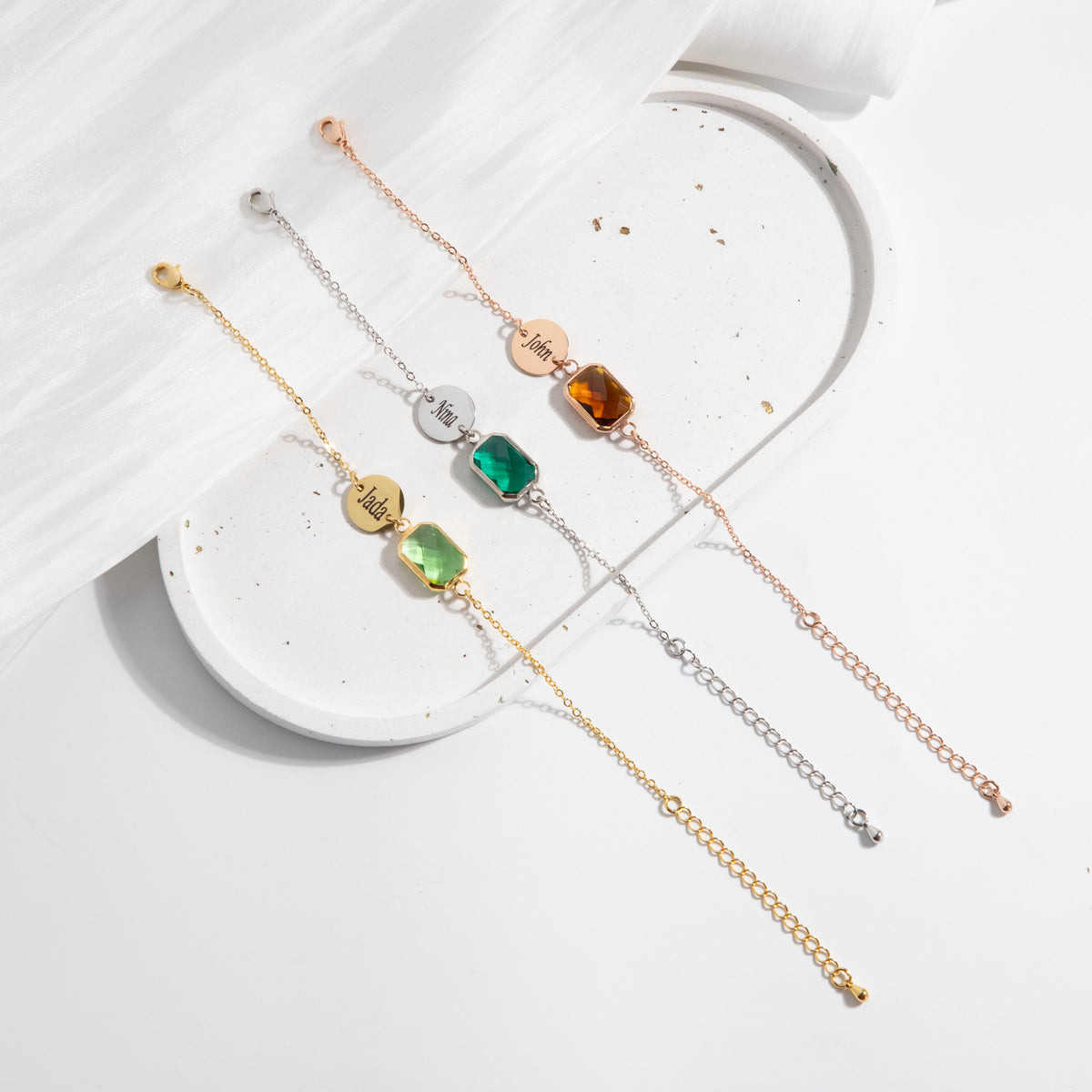 Three bracelets with engraved round discs and light green, green, and amber rectangular stones on a white display