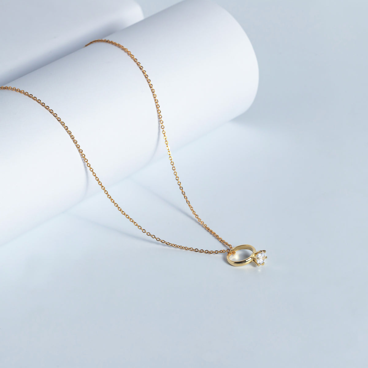Gold-tone necklace with a ring-shaped charm and clear stone displayed on a pale blue surface
