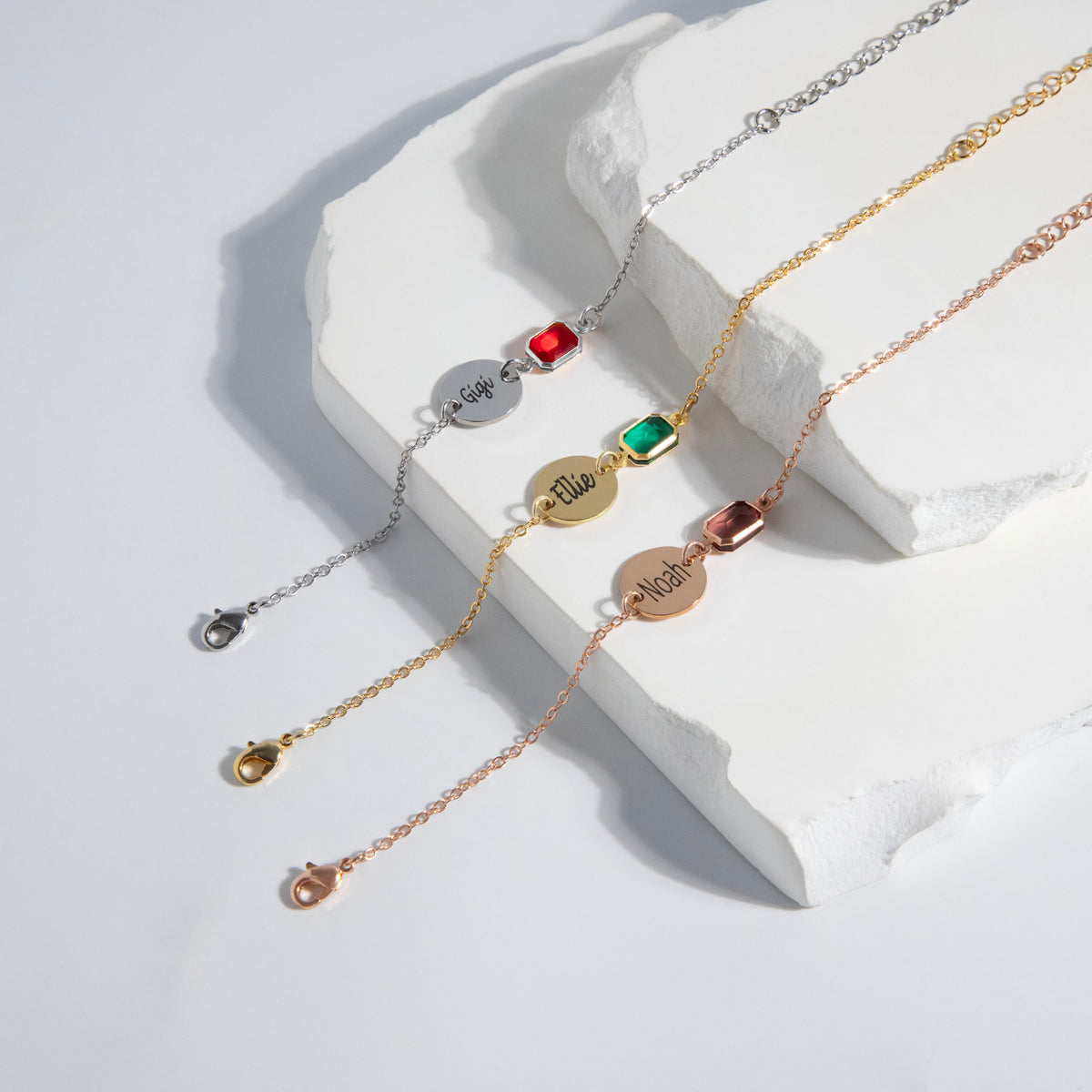 Three bracelets with engraved round discs and red, green, and brown rectangular stones on white sculptural surface
