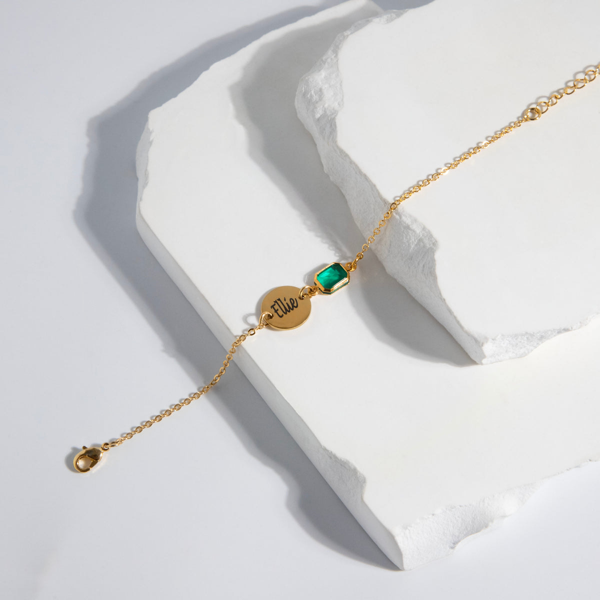Gold-tone bracelet with round disc engraved Ellie and green rectangular stone on a white sculptural surface
