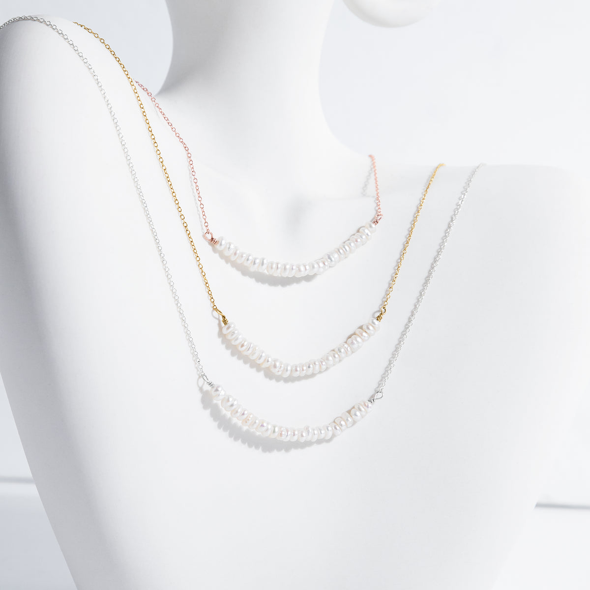 Three necklaces in silver-, gold-, and rose-gold tones with rows of small white pearls on a white display