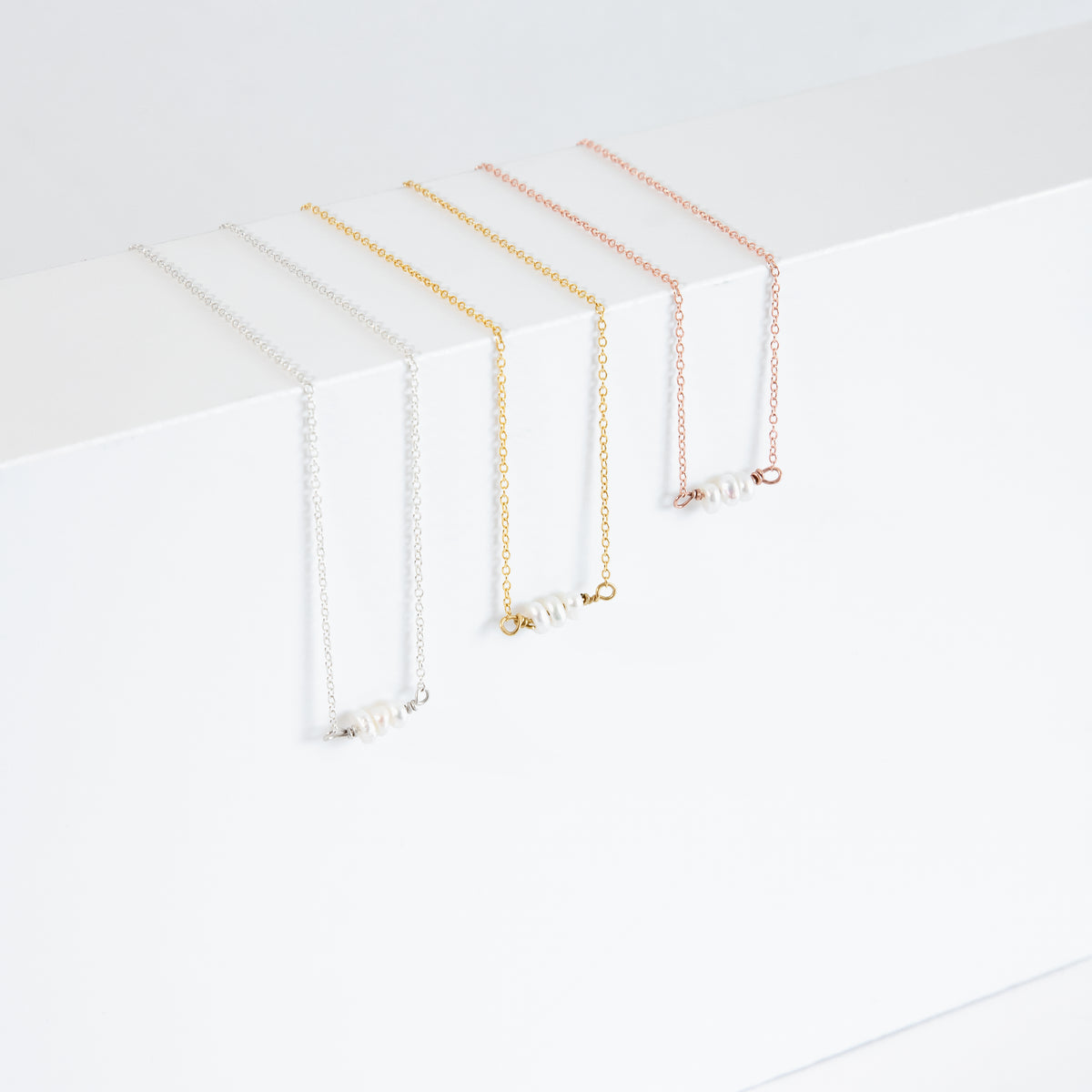 Three necklaces in silver-, gold-, and rose-gold tones with short rows of white oval pearls on a white display