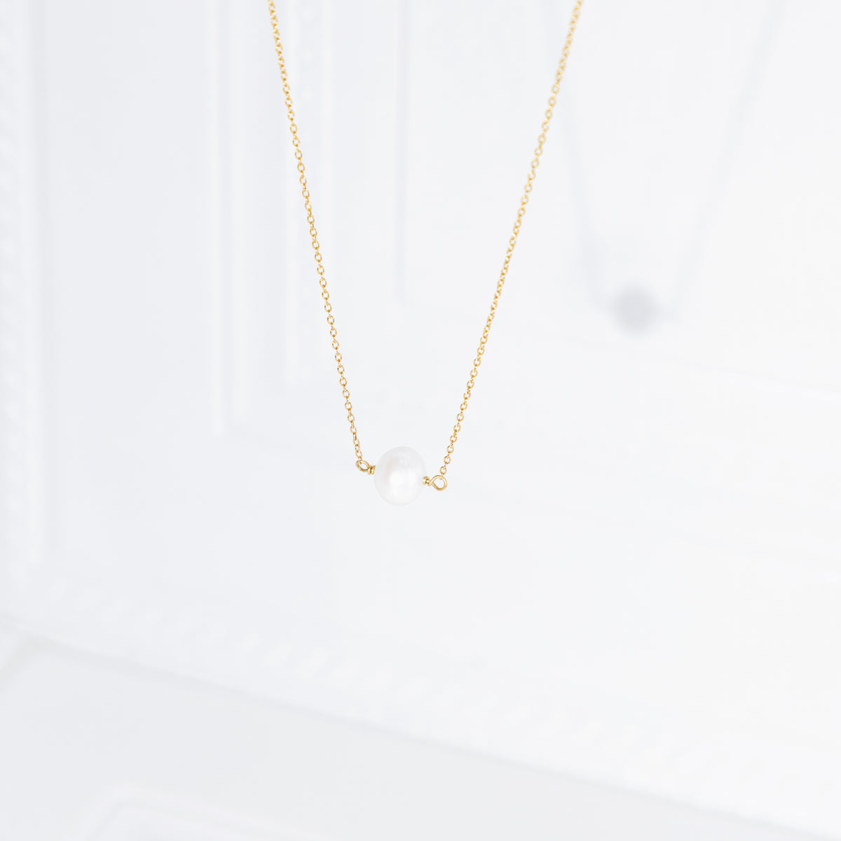 Gold-tone necklace with a single white pearl centered on the chain against a softly lit white background