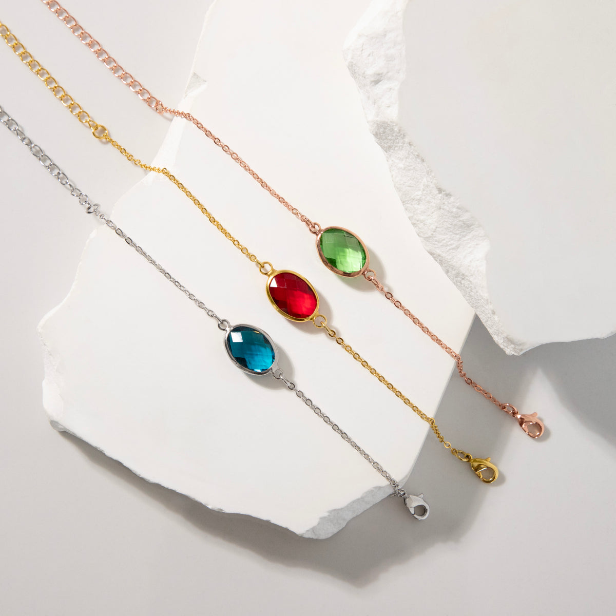 Three bracelets with blue, red, and green faceted oval stones arranged on a white sculptural surface