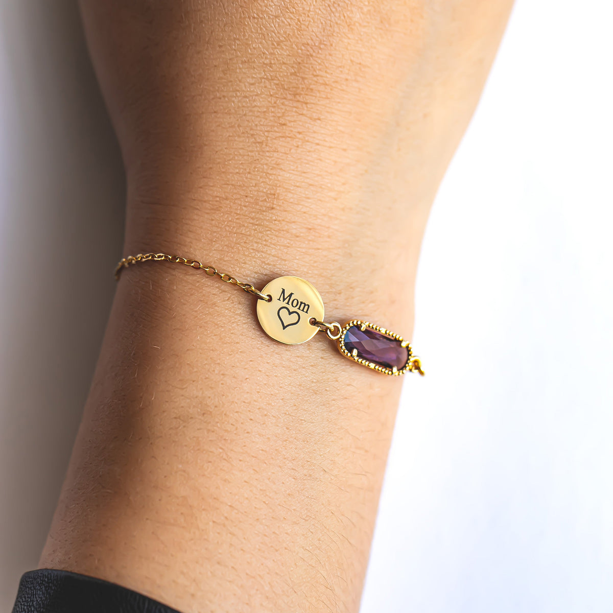 Gold-tone bracelet with round disc engraved Mom and heart beside a purple faceted rectangular stone worn on a wrist