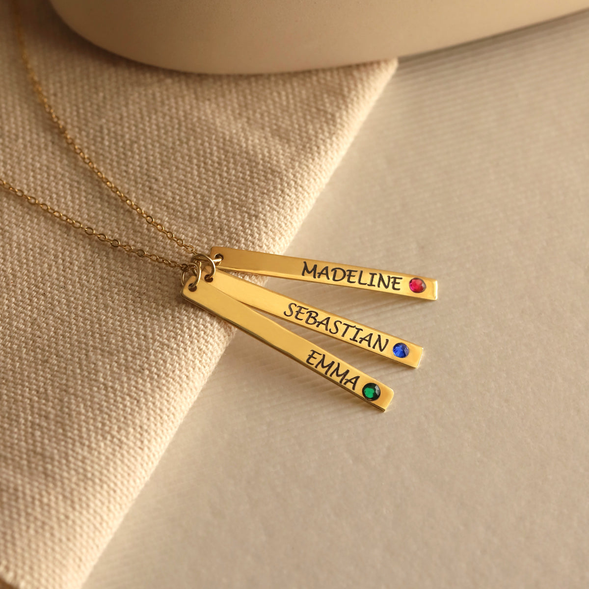 Gold-tone necklace with engraved bars reading MADELINE, SEBASTIAN, and EMMA with red, blue, and green stones