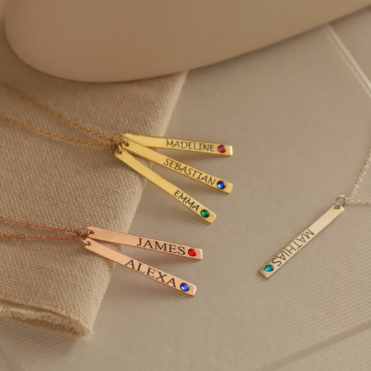 Gold-, rose-gold-, and silver-tone engraved bar necklaces with names and colored stones on beige fabric